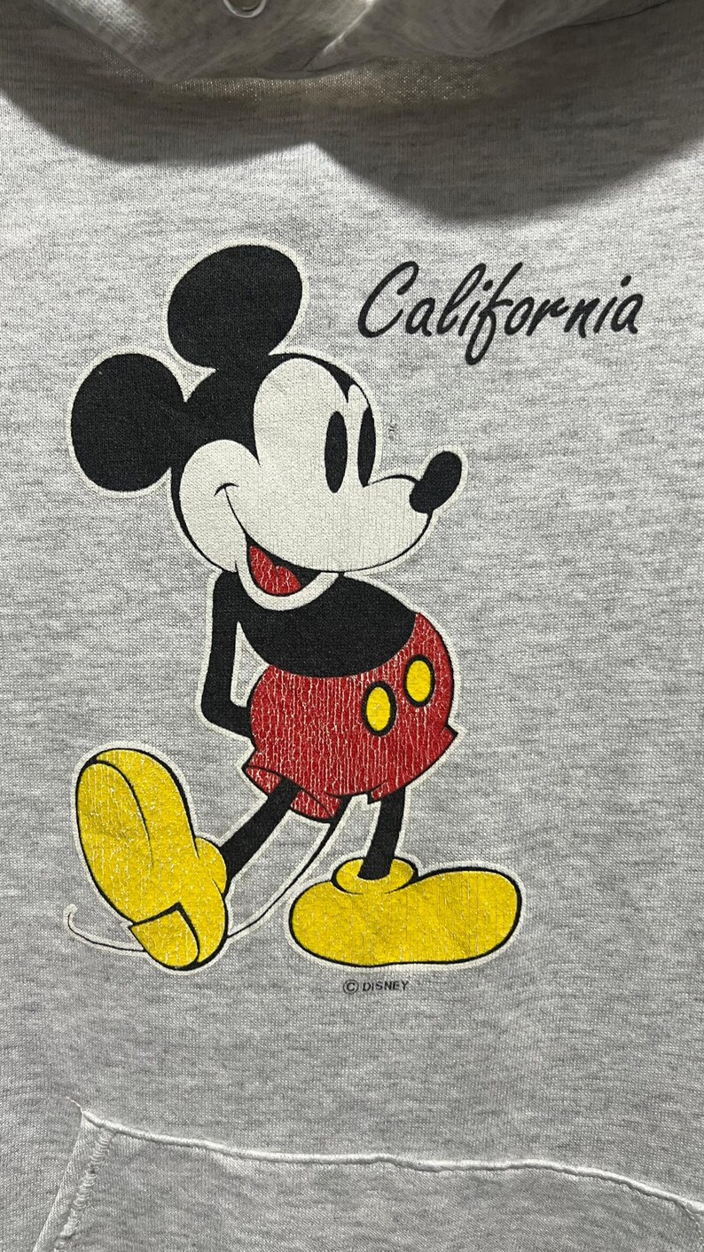 May include: A light grey hooded sweatshirt featuring a graphic of Mickey Mouse in black, white, red, and yellow. The character is positioned below the word California in black script. The Disney copyright symbol is printed below the character.