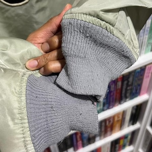 May include: A light sage green bomber jacket featuring the letters USA in large black felt-like applique with white trim. The jacket has ribbed grey knit cuffs at the sleeves and a smooth, slightly reflective synthetic fabric construction.