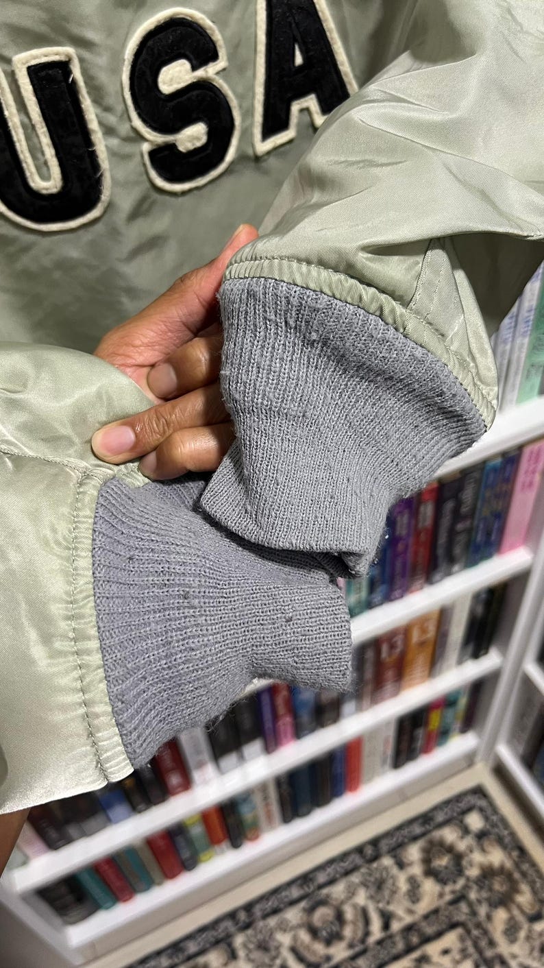 May include: A light sage green bomber jacket featuring the letters USA in large black felt-like applique with white trim. The jacket has ribbed grey knit cuffs at the sleeves and a smooth, slightly reflective synthetic fabric construction.