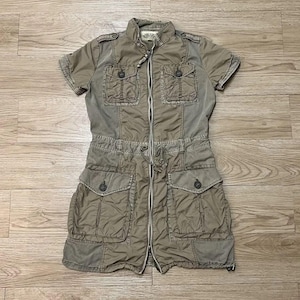 Puede incluir: Un vestido utilitario de color caqui con mangas cortas, cremallera frontal completa, cuello alzado, dos bolsillos abotonados en el pecho y dos bolsillos grandes abotonados en la cadera. La cintura incluye un cord&oacute;n ajustable.