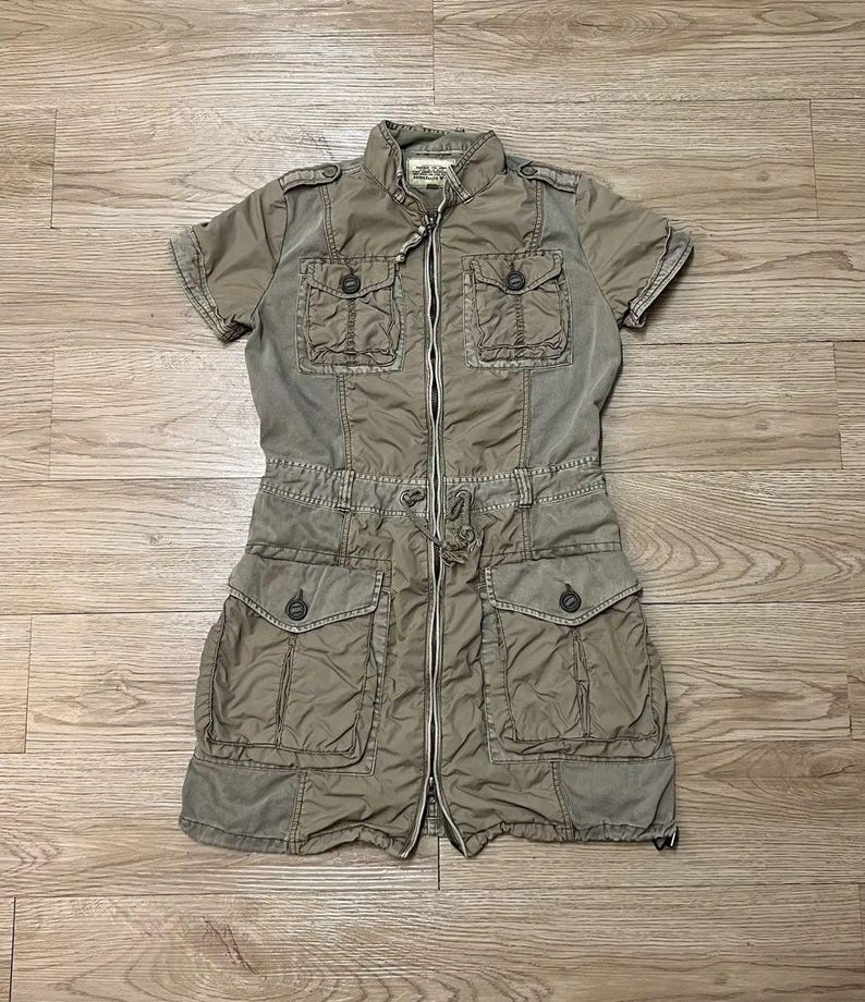 Puede incluir: Un vestido utilitario de color caqui con mangas cortas, cremallera frontal completa, cuello alzado, dos bolsillos abotonados en el pecho y dos bolsillos grandes abotonados en la cadera. La cintura incluye un cord&oacute;n ajustable.