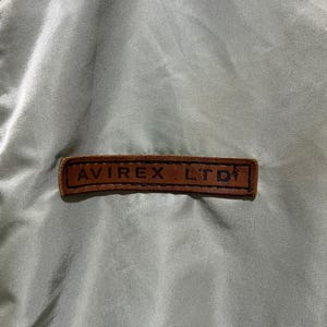 May include: A light olive green satin-finish jacket featuring a rectangular brown leather patch stitched onto the chest. The patch is embossed with the black text AVIREX LTD and includes a small graphic icon of a figure on the right side.