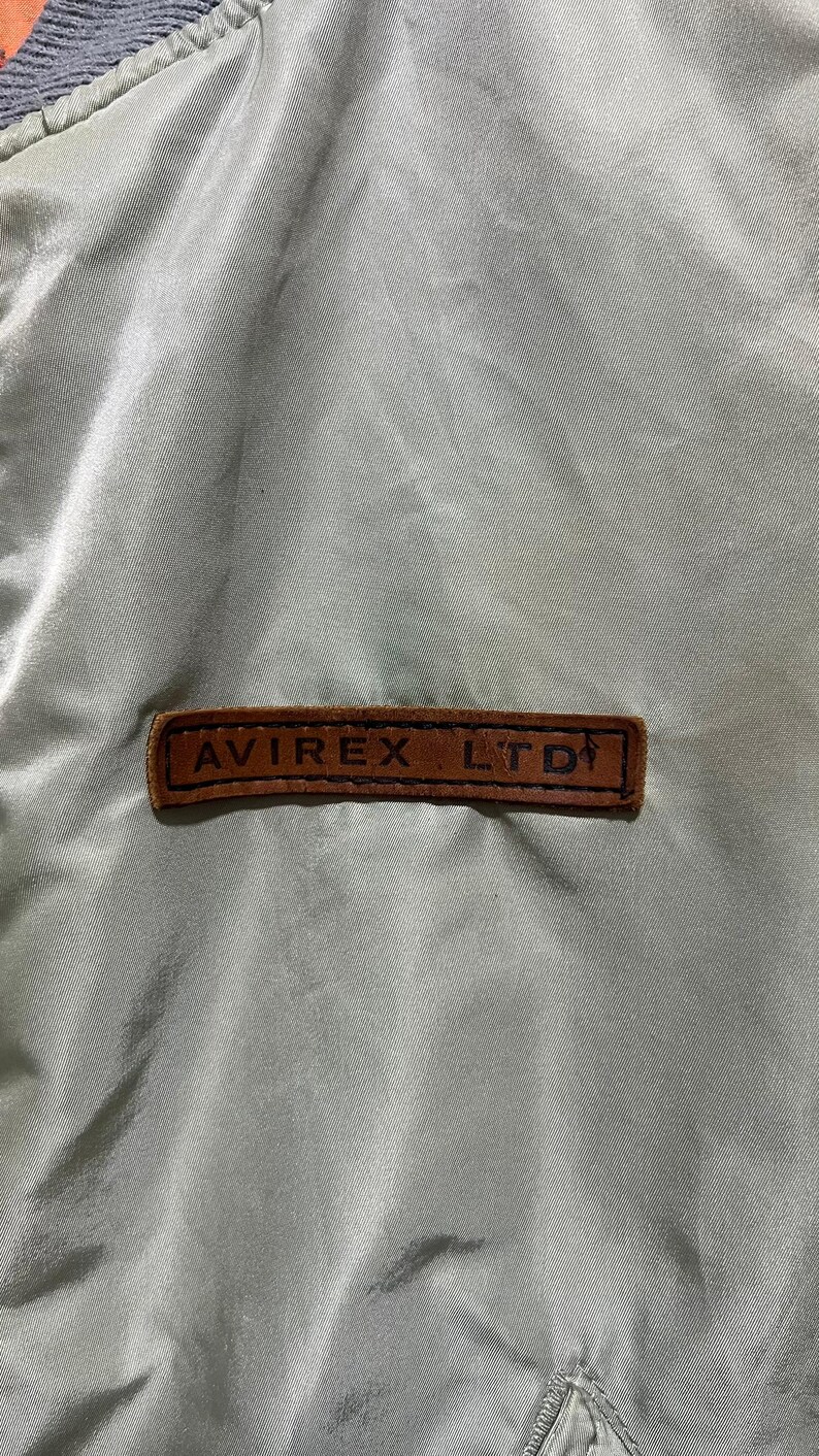 May include: A light olive green satin-finish jacket featuring a rectangular brown leather patch stitched onto the chest. The patch is embossed with the black text AVIREX LTD and includes a small graphic icon of a figure on the right side.