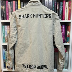 May include: A tan jacket with the words "SHARK HUNTERS" printed across the back in black block letters. Below the text is "75 LRSP SQDRN." The jacket is hanging on a hanger in front of a bookshelf.