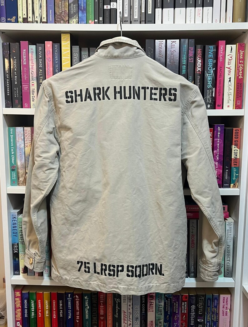 May include: A tan jacket with the words "SHARK HUNTERS" printed across the back in black block letters. Below the text is "75 LRSP SQDRN." The jacket is hanging on a hanger in front of a bookshelf.