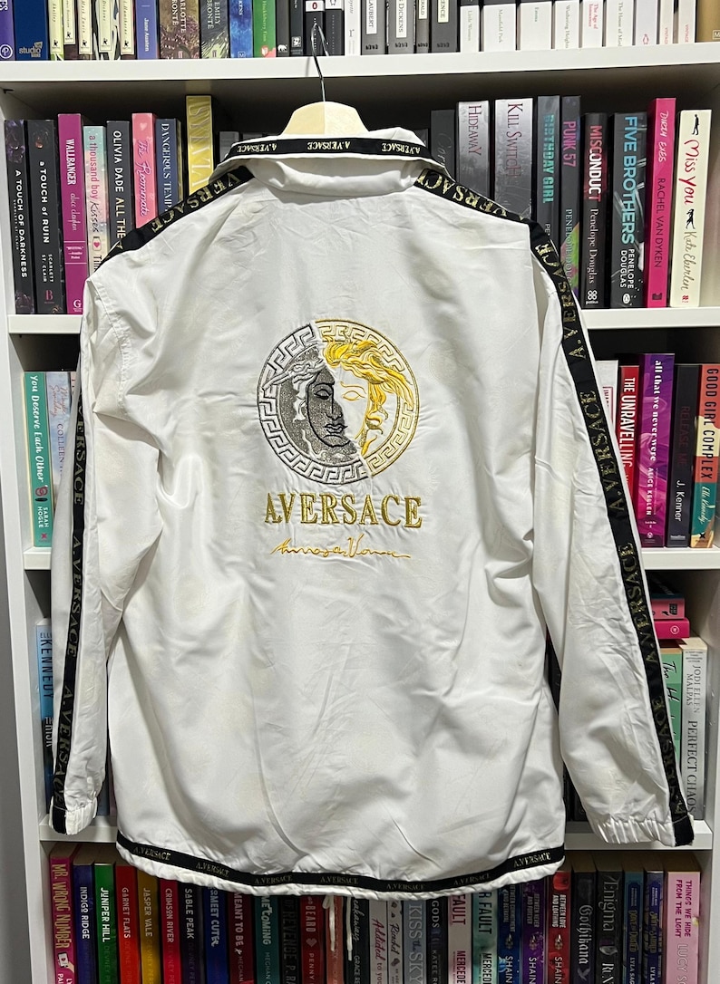 May include: A white windbreaker jacket featuring black stripes along the sleeves and hem with the text A.VERSACE. The back displays a large circular embroidered logo split between silver and gold tones, with the text AVERSACE embroidered below.