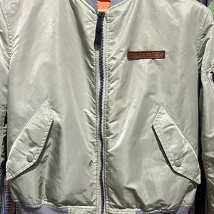 May include: A sage green bomber jacket featuring a ribbed grey collar and hem, a central black zipper, and two front pockets with snap closures. A brown rectangular patch with the text AVIREX LTD is stitched on the upper left chest area.
