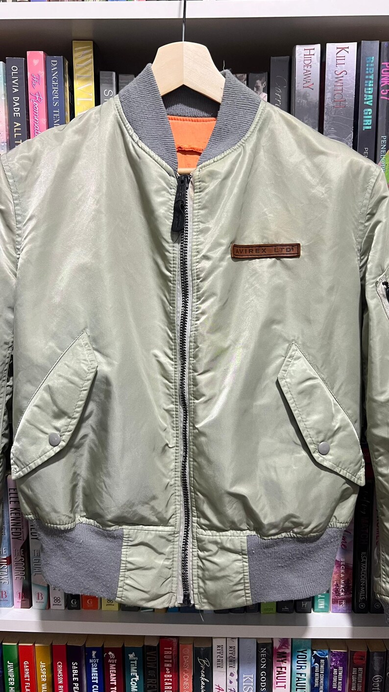 May include: A sage green bomber jacket featuring a ribbed grey collar and hem, a central black zipper, and two front pockets with snap closures. A brown rectangular patch with the text AVIREX LTD is stitched on the upper left chest area.