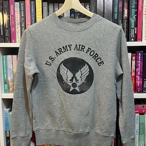 May include: A gray crewneck sweatshirt with the text "U.S. ARMY AIR FORCE" in black, encircling a graphic of wings and a star. The sweatshirt is displayed on a wooden hanger against a backdrop of bookshelves filled with books.