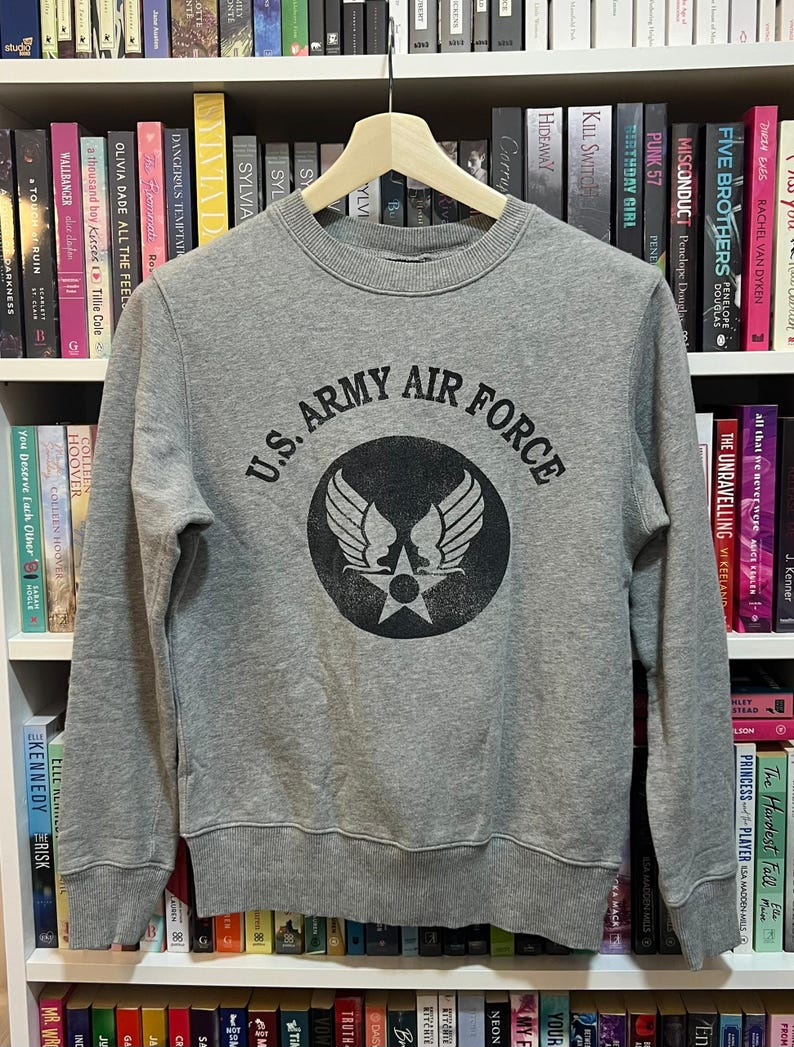 May include: A gray crewneck sweatshirt with the text "U.S. ARMY AIR FORCE" in black, encircling a graphic of wings and a star. The sweatshirt is displayed on a wooden hanger against a backdrop of bookshelves filled with books.
