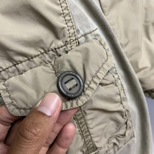 Puede incluir: Una chaqueta de color beige que presenta un bolsillo con solapa y un bot&oacute;n circular de color marr&oacute;n oscuro. El bot&oacute;n tiene grabado el texto AVIREX. La chaqueta est&aacute; confeccionada con una combinaci&oacute;n de tejido sint&eacute;tico liso y un panel de material tejido texturizado.