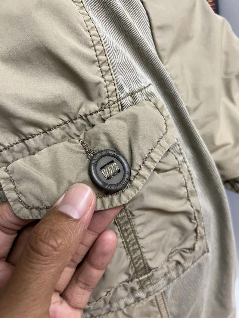 Puede incluir: Una chaqueta de color beige que presenta un bolsillo con solapa y un bot&oacute;n circular de color marr&oacute;n oscuro. El bot&oacute;n tiene grabado el texto AVIREX. La chaqueta est&aacute; confeccionada con una combinaci&oacute;n de tejido sint&eacute;tico liso y un panel de material tejido texturizado.