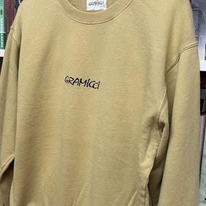 May include: A tan Gramicci sweatshirt with a crew neck and long sleeves. The brand name is embroidered in black script across the chest. The sweatshirt is hanging on a wooden hanger in front of a bookshelf.