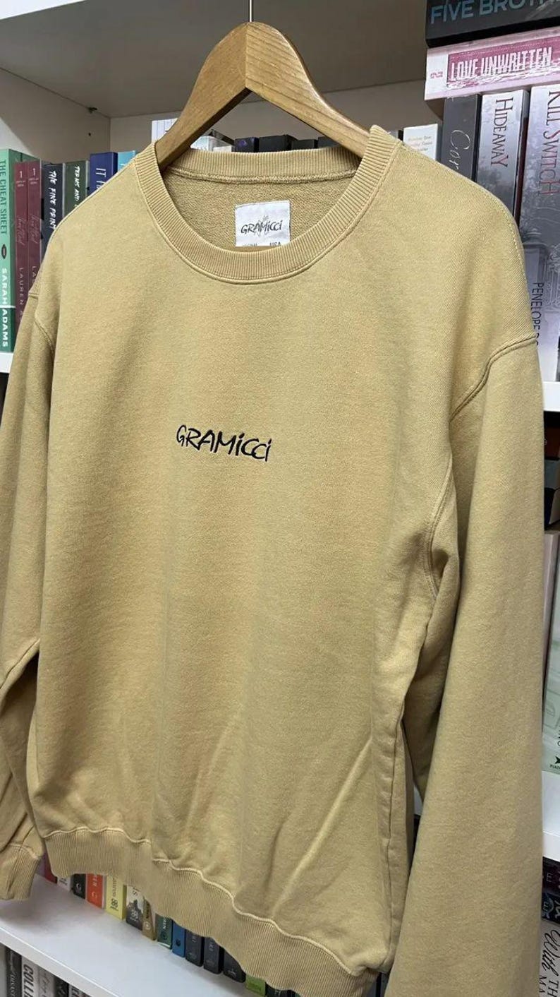 May include: A tan Gramicci sweatshirt with a crew neck and long sleeves. The brand name is embroidered in black script across the chest. The sweatshirt is hanging on a wooden hanger in front of a bookshelf.