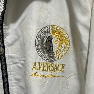 May include: A white zip-up jacket featuring a tonal repeating pattern and a central embroidered circular logo. The logo is split between silver and gold, depicting a face, with the text A.VERSACE embroidered in gold thread below it.