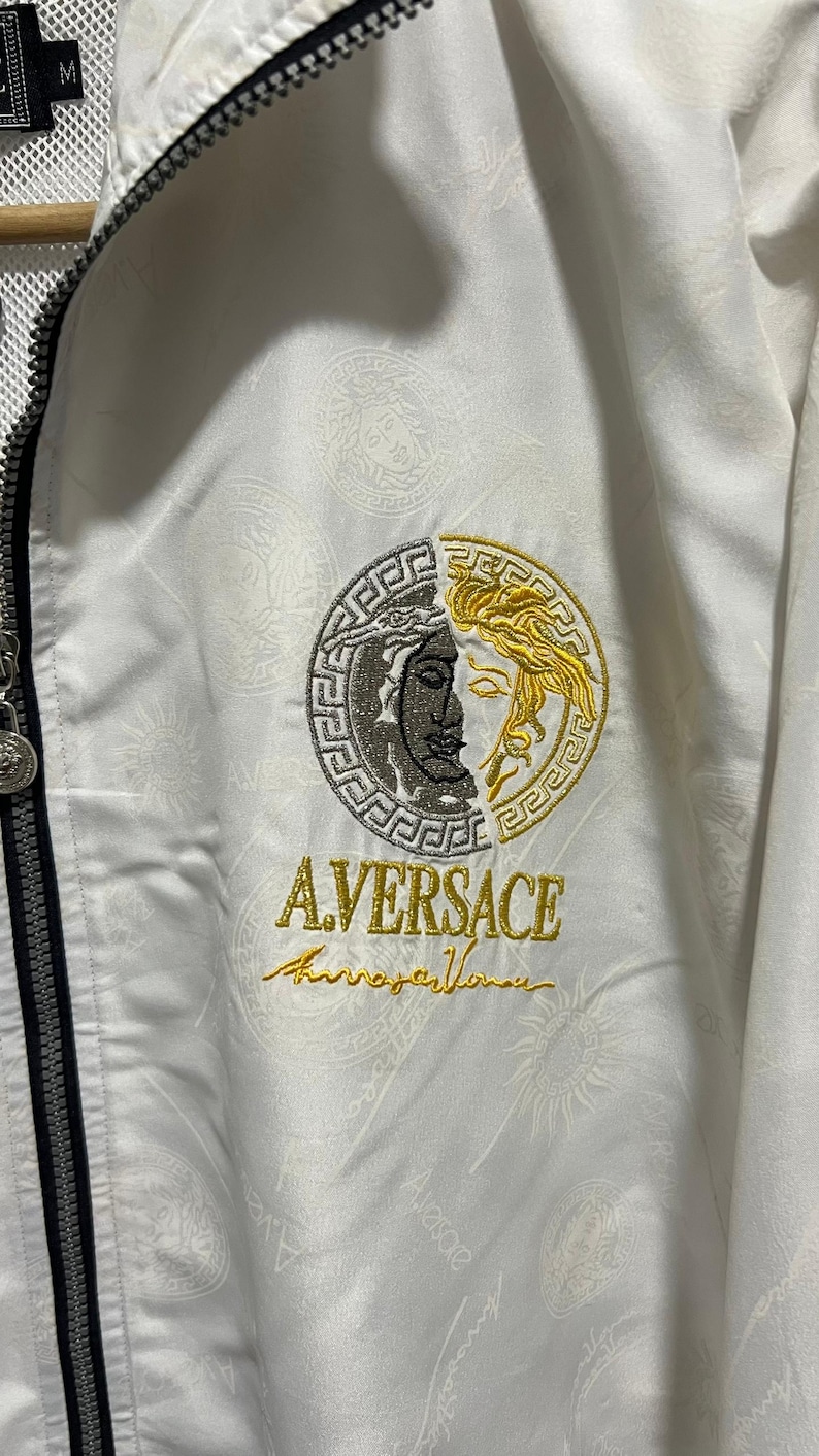 May include: A white zip-up jacket featuring a tonal repeating pattern and a central embroidered circular logo. The logo is split between silver and gold, depicting a face, with the text A.VERSACE embroidered in gold thread below it.