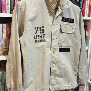 May include: Tan military-style jacket with button closures and a collared neckline. Features the text "75 LRSP SOORN" on the front and a "U.S. NAVY" patch. The jacket has two front pockets and is hanging on a wooden hanger.