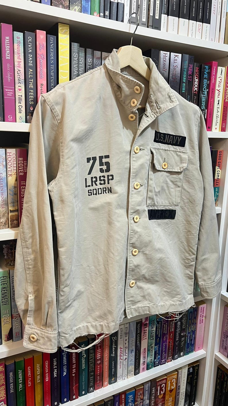 May include: Tan military-style jacket with button closures and a collared neckline. Features the text "75 LRSP SOORN" on the front and a "U.S. NAVY" patch. The jacket has two front pockets and is hanging on a wooden hanger.