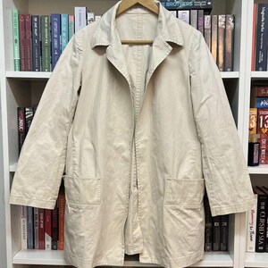 Puede incluir: Un abrigo de color beige claro con mangas largas, cuello cl&aacute;sico, cierre frontal con botones y dos grandes bolsillos rectangulares de parche situados en la parte inferior delantera. La prenda est&aacute; confeccionada en tela tejida lisa.