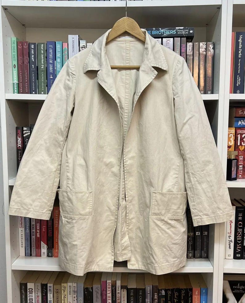 Puede incluir: Un abrigo de color beige claro con mangas largas, cuello cl&aacute;sico, cierre frontal con botones y dos grandes bolsillos rectangulares de parche situados en la parte inferior delantera. La prenda est&aacute; confeccionada en tela tejida lisa.