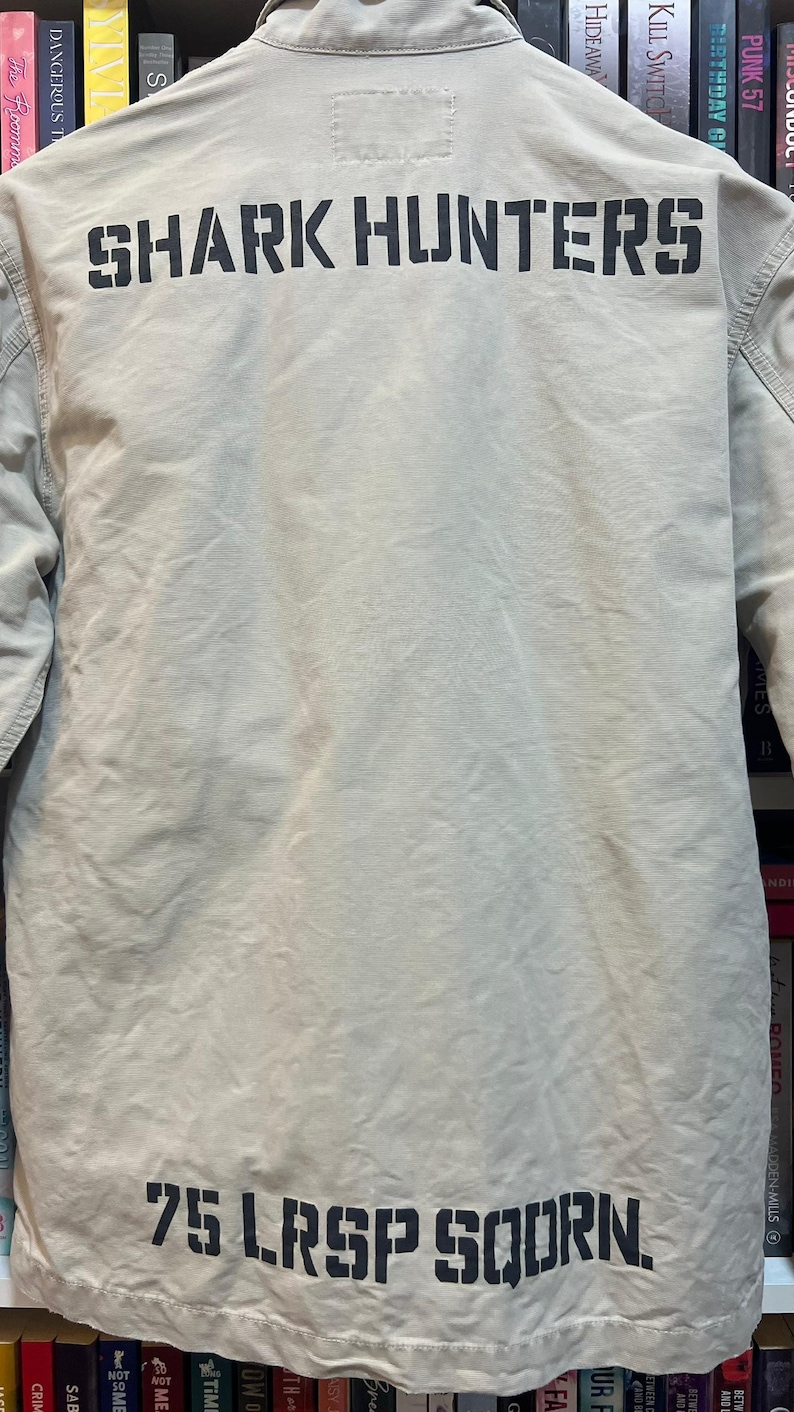 May include: Tan jacket with the words "SHARK HUNTERS" printed across the back in black block letters. Below the text is "75 LRSP SQDRN." The jacket is made of a light-colored fabric.