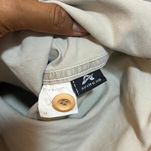May include: Close-up of a tan garment with a button and a tag. The tag features the brand name AVIREX in white text on a black background. The garment is a light beige color.