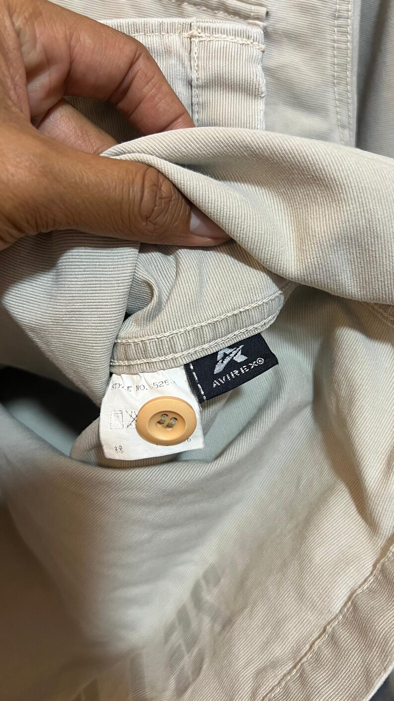 May include: Close-up of a tan garment with a button and a tag. The tag features the brand name AVIREX in white text on a black background. The garment is a light beige color.