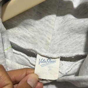 May include: A light grey hooded sweatshirt featuring a white interior neck label with the blue text Velva Sheen, the size M, and the text OVER FOR CARE MADE IN THE U.S.A. The hoodie is displayed on a wooden hanger with a partial black graphic visible.