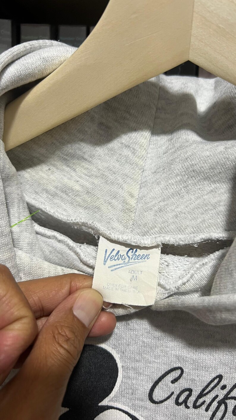 May include: A light grey hooded sweatshirt featuring a white interior neck label with the blue text Velva Sheen, the size M, and the text OVER FOR CARE MADE IN THE U.S.A. The hoodie is displayed on a wooden hanger with a partial black graphic visible.