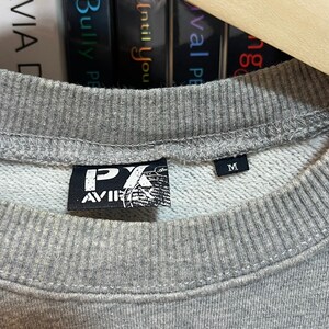 May include: Close-up of a gray sweatshirt with a crew neck. The sweatshirt has a black label with white text and a graphic. The size tag reads "M". The sweatshirt is hanging on a wooden hanger.