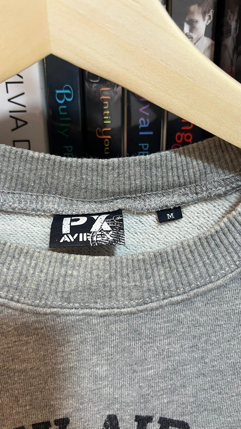 May include: Close-up of a gray sweatshirt with a crew neck. The sweatshirt has a black label with white text and a graphic. The size tag reads "M". The sweatshirt is hanging on a wooden hanger.