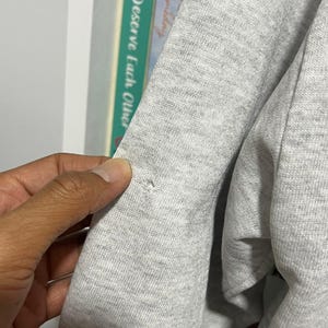 May include: A light grey heathered sweatshirt featuring a small, circular hole in the fabric on the sleeve. The garment is made of a soft, textured knit material and displays a visible imperfection in the weave of the grey fabric.