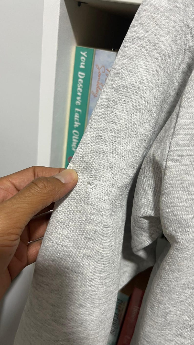 May include: A light grey heathered sweatshirt featuring a small, circular hole in the fabric on the sleeve. The garment is made of a soft, textured knit material and displays a visible imperfection in the weave of the grey fabric.