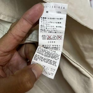 Puede incluir: Una etiqueta de cuidado de tela blanca cosida en una prenda beige. La etiqueta presenta texto en negro que incluye 'YM-J15-014', '100% algod&oacute;n' y varios s&iacute;mbolos de lavado. Incluye instrucciones en japon&eacute;s, franc&eacute;s e ingl&eacute;s sobre el lavado.