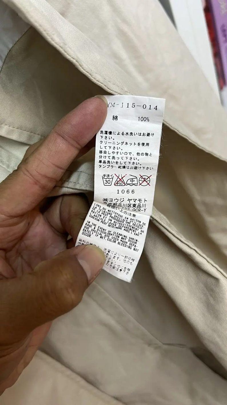 Puede incluir: Una etiqueta de cuidado de tela blanca cosida en una prenda beige. La etiqueta presenta texto en negro que incluye 'YM-J15-014', '100% algod&oacute;n' y varios s&iacute;mbolos de lavado. Incluye instrucciones en japon&eacute;s, franc&eacute;s e ingl&eacute;s sobre el lavado.