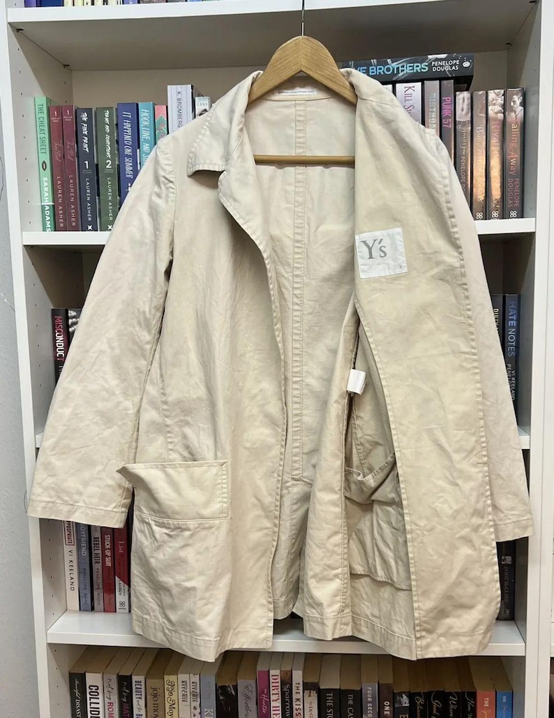 Puede incluir: Una chaqueta de algod&oacute;n beige colgada en una percha de madera. La chaqueta presenta un cuello ancho, mangas largas y dos grandes bolsillos frontales. Una etiqueta blanca con el texto Y's est&aacute; cosida en el interior del pecho izquierdo.