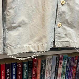 May include: Close-up of a light beige corduroy jacket with visible buttons and frayed edges. The jacket is partially unbuttoned, revealing a glimpse of a bookshelf filled with colorful books.