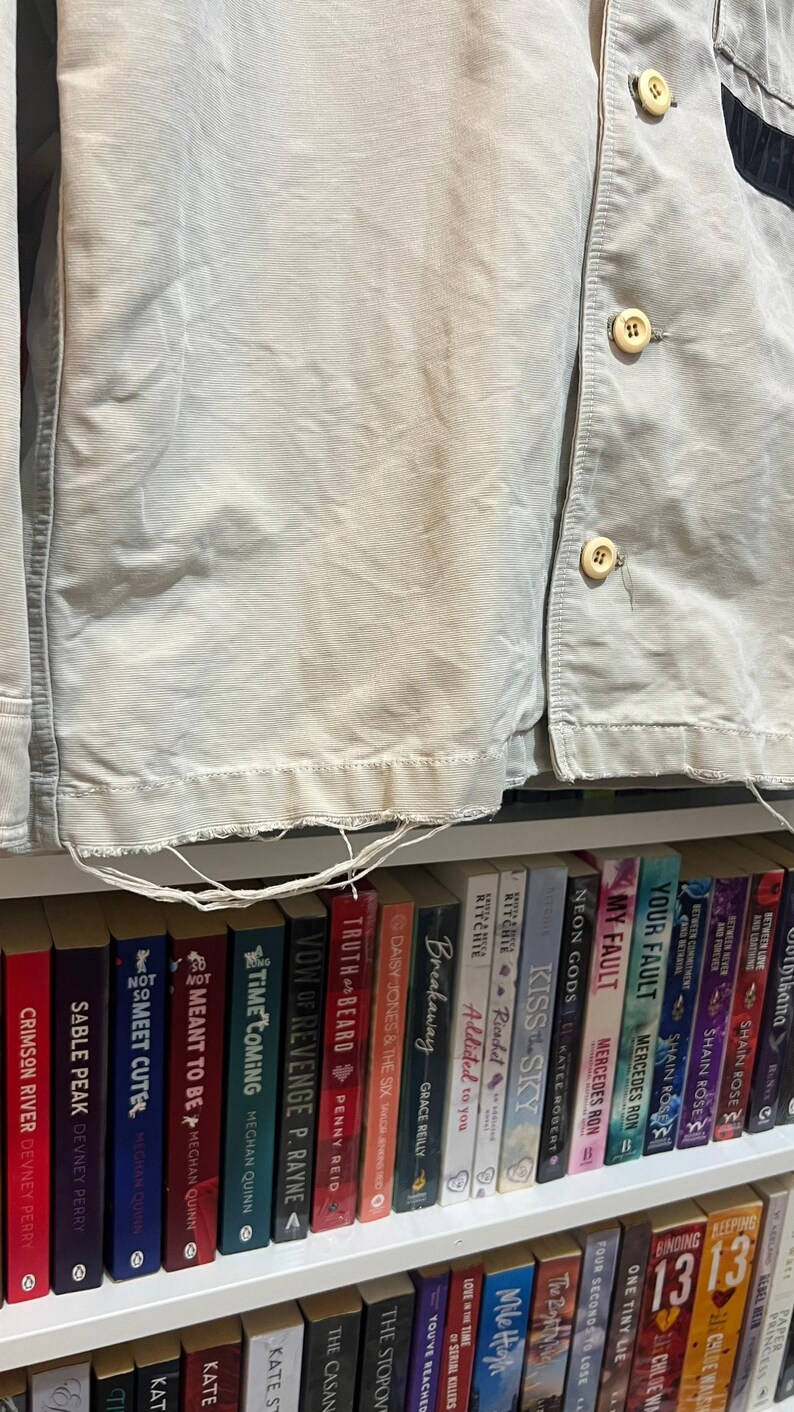 May include: Close-up of a light beige corduroy jacket with visible buttons and frayed edges. The jacket is partially unbuttoned, revealing a glimpse of a bookshelf filled with colorful books.