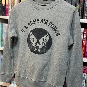 May include: A gray sweatshirt with the text "U.S. ARMY AIR FORCE" in black, surrounding a circular emblem with wings and a star. The sweatshirt is hanging on a wooden hanger.