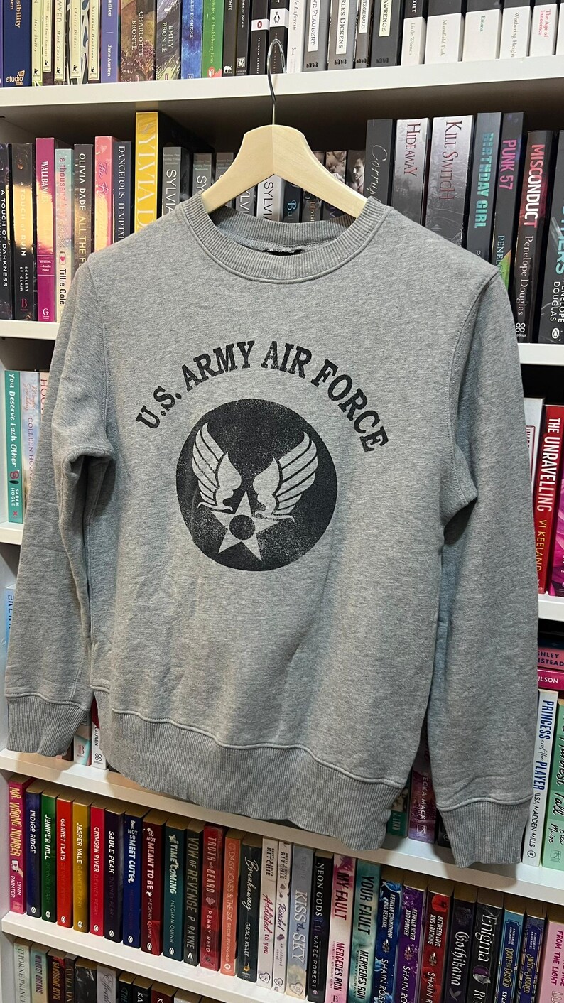 May include: A gray sweatshirt with the text "U.S. ARMY AIR FORCE" in black, surrounding a circular emblem with wings and a star. The sweatshirt is hanging on a wooden hanger.