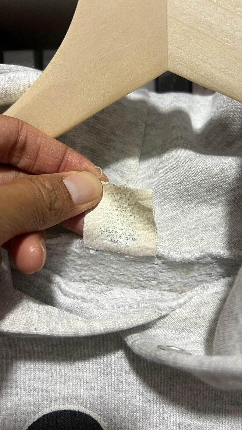 May include: A light grey hooded sweatshirt featuring a white fabric care label sewn into the inner neckline. The label includes printed text with washing instructions: Machine wash warm, wash inside out with like colours, do not bleach, tumble dry low, do not iron design, do not dry clean, and RN 61625.