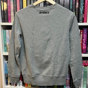 May include: A heather gray sweatshirt with the brand "AVIREX" printed in black on the upper back. The sweatshirt is hanging on a wooden hanger in front of a bookshelf filled with books.