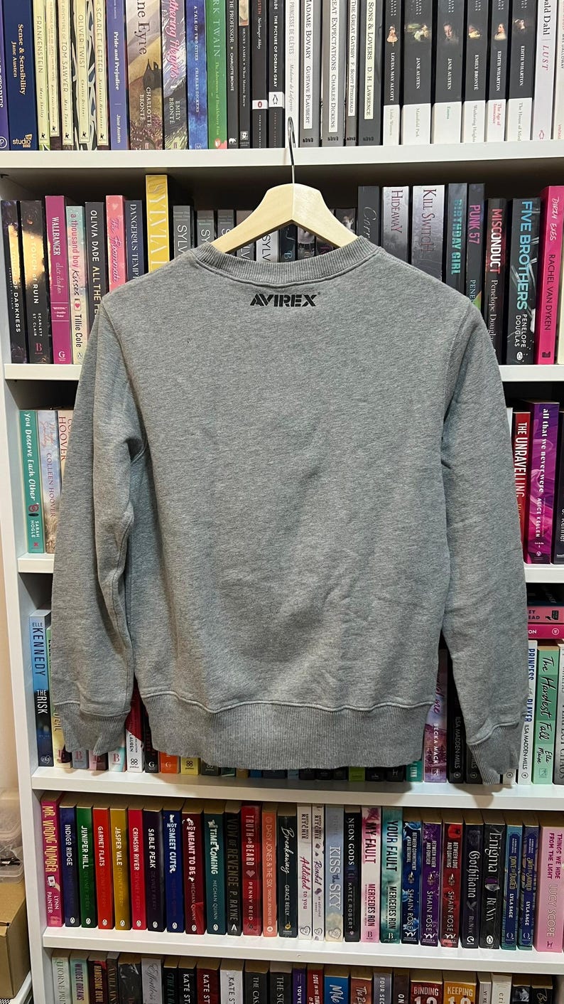 May include: A heather gray sweatshirt with the brand "AVIREX" printed in black on the upper back. The sweatshirt is hanging on a wooden hanger in front of a bookshelf filled with books.