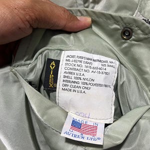 May include: A sage green nylon flight jacket with a white interior label reading JACKET, FLYER'S MAN INTERMEDIATE, MA-1, MIL-J-8279E (USAF), SIZE SMALL, STOCK NO. 1615-522-6014, CONTRACT NO. AV-15-37801, AVIREX U.S.A., SHELL 100% NYLON, INTERLINING: 100% POLYESTER FIBERFILL, DRY CLEAN ONLY, MADE IN U.S.A. and a small tag with an American flag and AVIREX LTD.