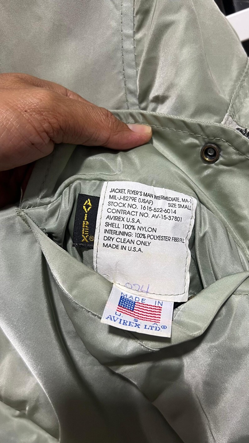 May include: A sage green nylon flight jacket with a white interior label reading JACKET, FLYER'S MAN INTERMEDIATE, MA-1, MIL-J-8279E (USAF), SIZE SMALL, STOCK NO. 1615-522-6014, CONTRACT NO. AV-15-37801, AVIREX U.S.A., SHELL 100% NYLON, INTERLINING: 100% POLYESTER FIBERFILL, DRY CLEAN ONLY, MADE IN U.S.A. and a small tag with an American flag and AVIREX LTD.