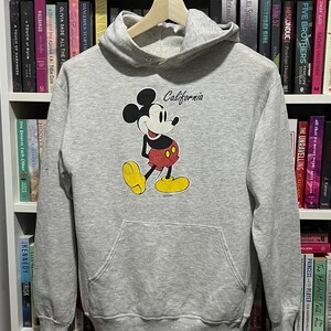 May include: A light grey hooded sweatshirt featuring a graphic of Mickey Mouse in red shorts and yellow shoes. The word California is printed in black script above the character. The garment includes a front kangaroo pocket and long sleeves.