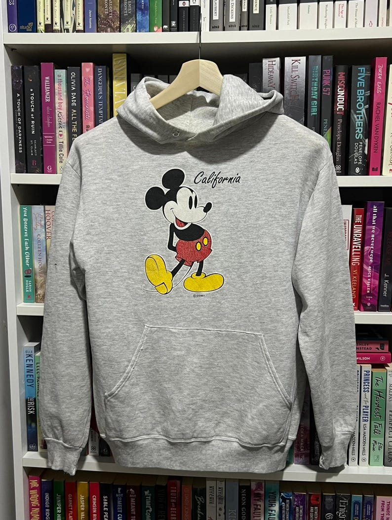 May include: A light grey hooded sweatshirt featuring a graphic of Mickey Mouse in red shorts and yellow shoes. The word California is printed in black script above the character. The garment includes a front kangaroo pocket and long sleeves.