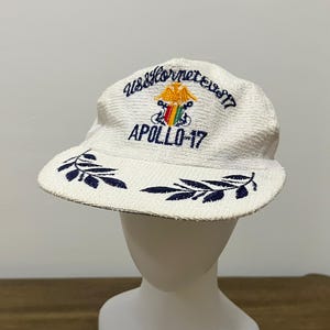 Op de afbeelding: Een witte, getextureerde honkbalpet met donkerblauw borduurwerk met de tekst USS Hornet CVS-17 en APOLLO-17. De voorkant toont een geel adelaarembleem boven een schild met rode, gele en groene strepen, geflankeerd door donkerblauwe bladmotieven.