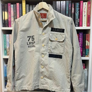 May include: A tan U.S. Navy jacket with a button-up front, two chest pockets, and patches. The jacket features the number "75" and the text "LRSP SQDRN" printed on the front. The jacket is hanging on a wooden hanger.