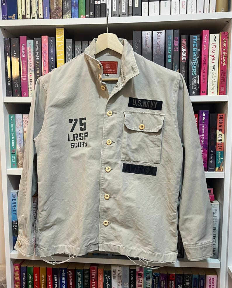 May include: A tan U.S. Navy jacket with a button-up front, two chest pockets, and patches. The jacket features the number "75" and the text "LRSP SQDRN" printed on the front. The jacket is hanging on a wooden hanger.